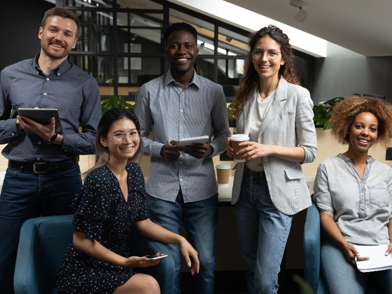 Five smiling people, casually dressed, stand and sit together in a modern office, holding tablets and coffee.