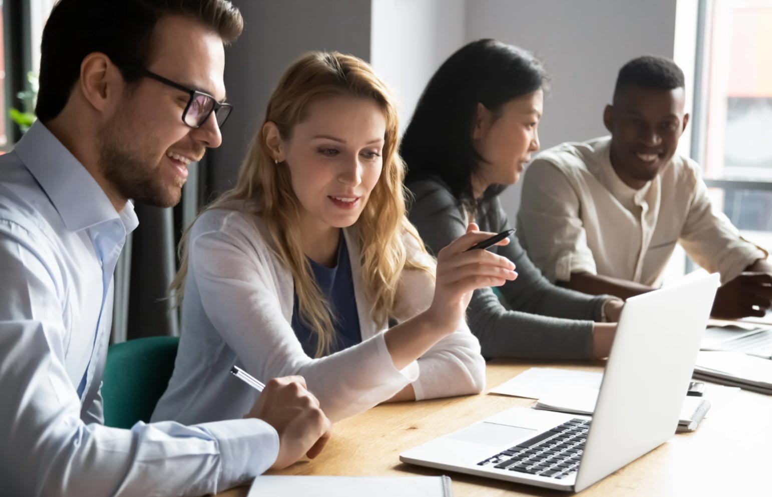 Four people sit at a table, collaborating and smiling while looking at a laptop computer.