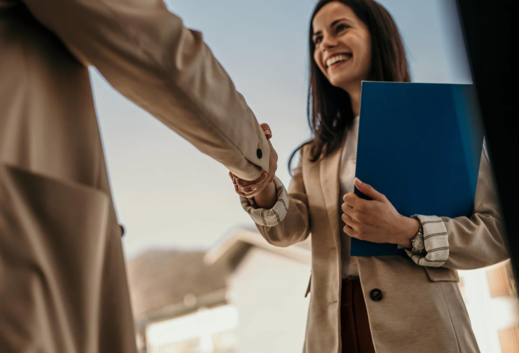 Smiling woman holding a blue folder shakes hands with another person outdoors.