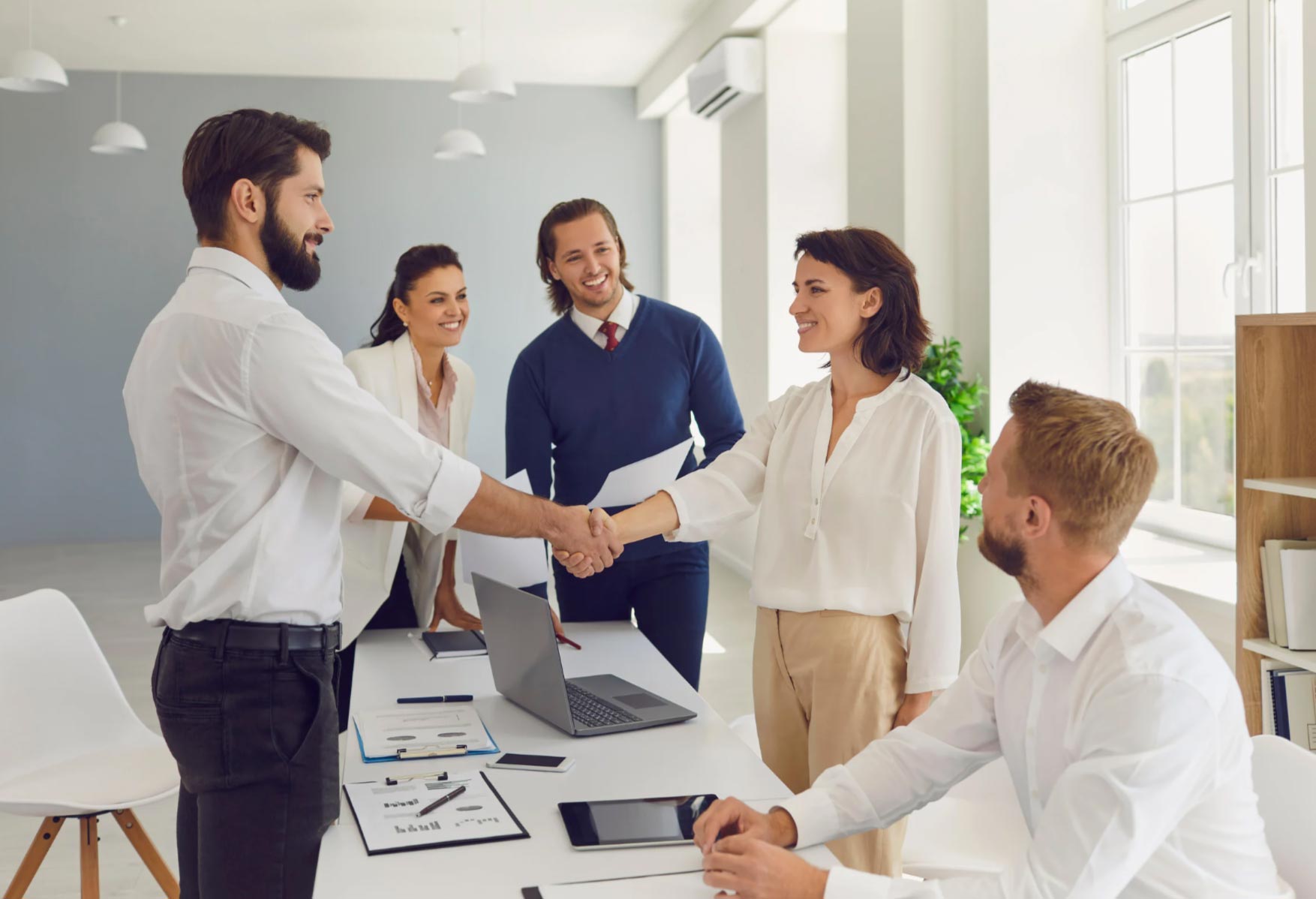 Two business people shake hands at a meeting table, while three colleagues watch and smile in a bright office.