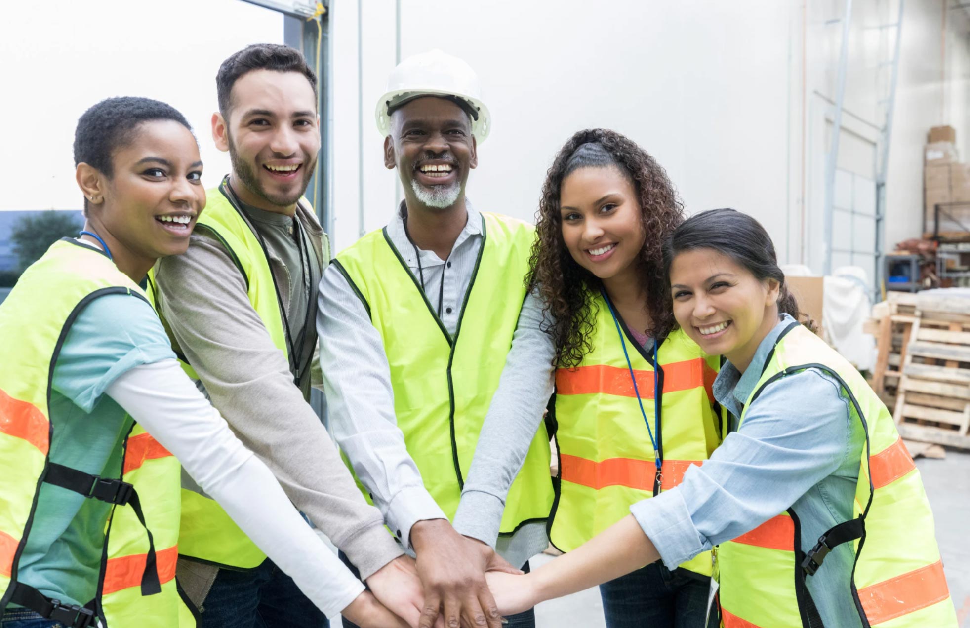 Five warehouse workers in safety vests smiling and stacking hands together in teamwork inside a warehouse.