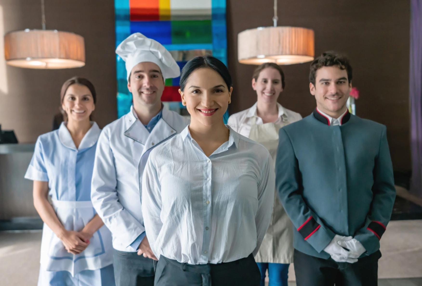 Five hospitality staff members in uniforms standing and smiling indoors, facing the camera.