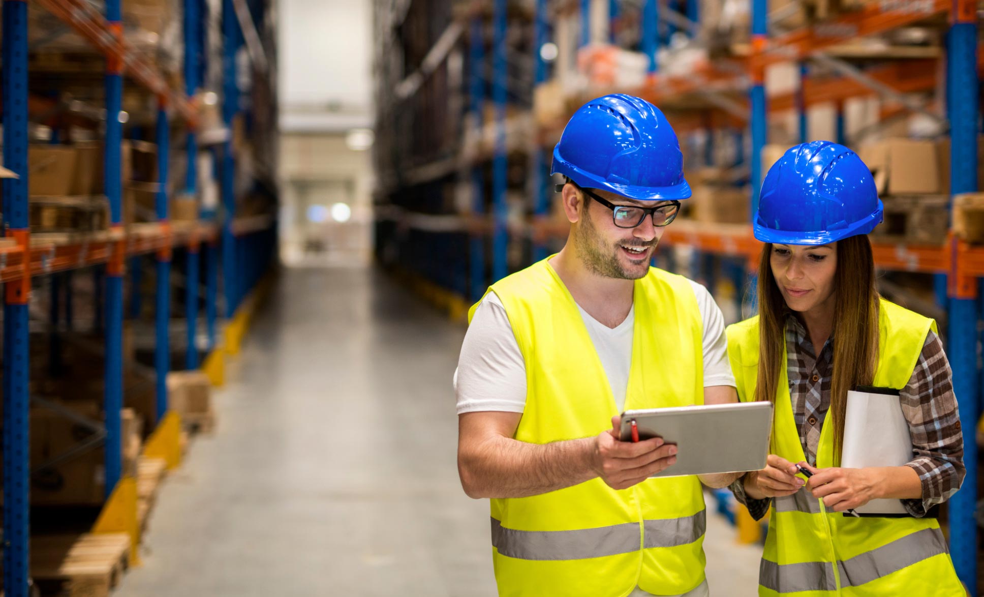 Two warehouse workers in safety vests and helmets looking at a tablet, surrounded by shelves of boxes.