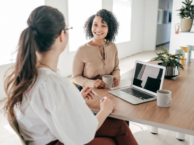 Two women smiling and talking at a desk with a laptop and coffee mugs in a bright office.