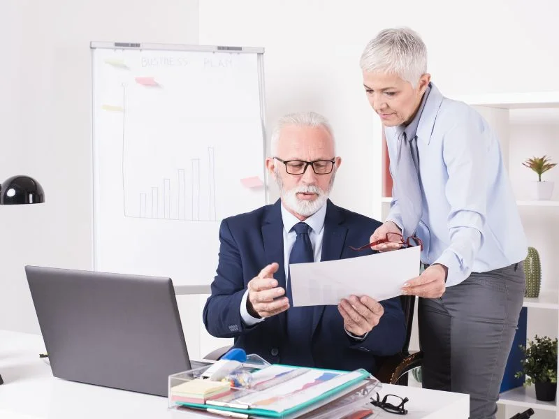 Two businesspeople in an office review a document together in front of a laptop and a flip chart.
