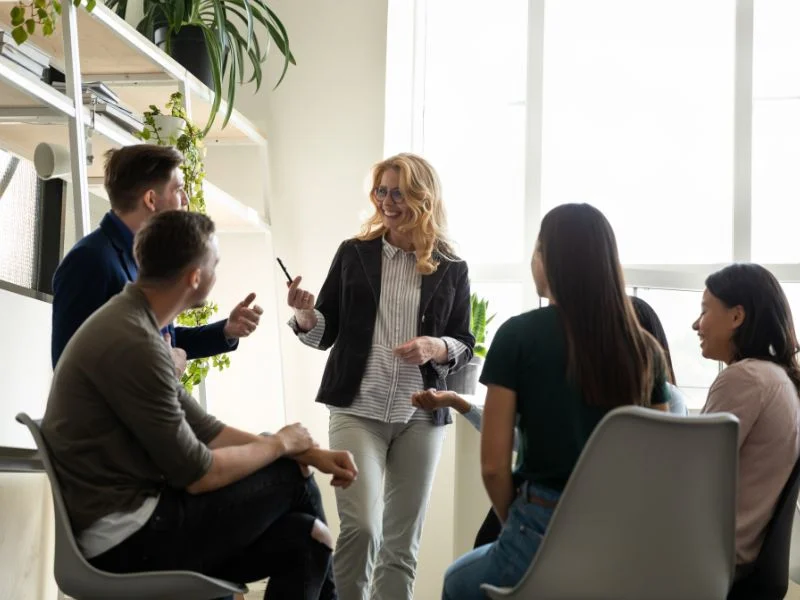 A woman stands and gestures while speaking to four seated people in a bright, modern office space.