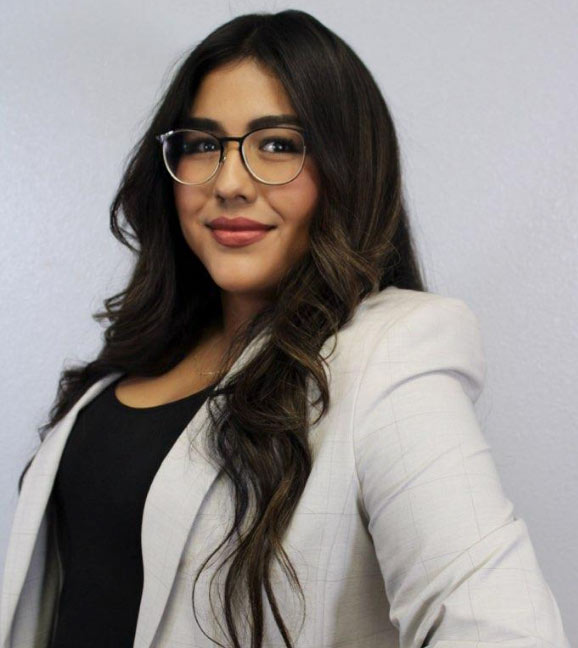 Woman with long dark hair, glasses, and a light blazer, smiling in front of a plain light background.