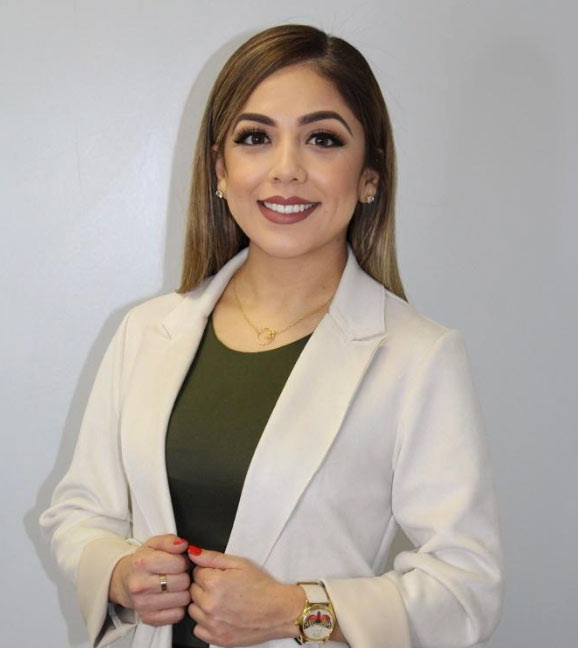 Woman with long hair in a white blazer and green top, smiling in front of a plain light gray background.